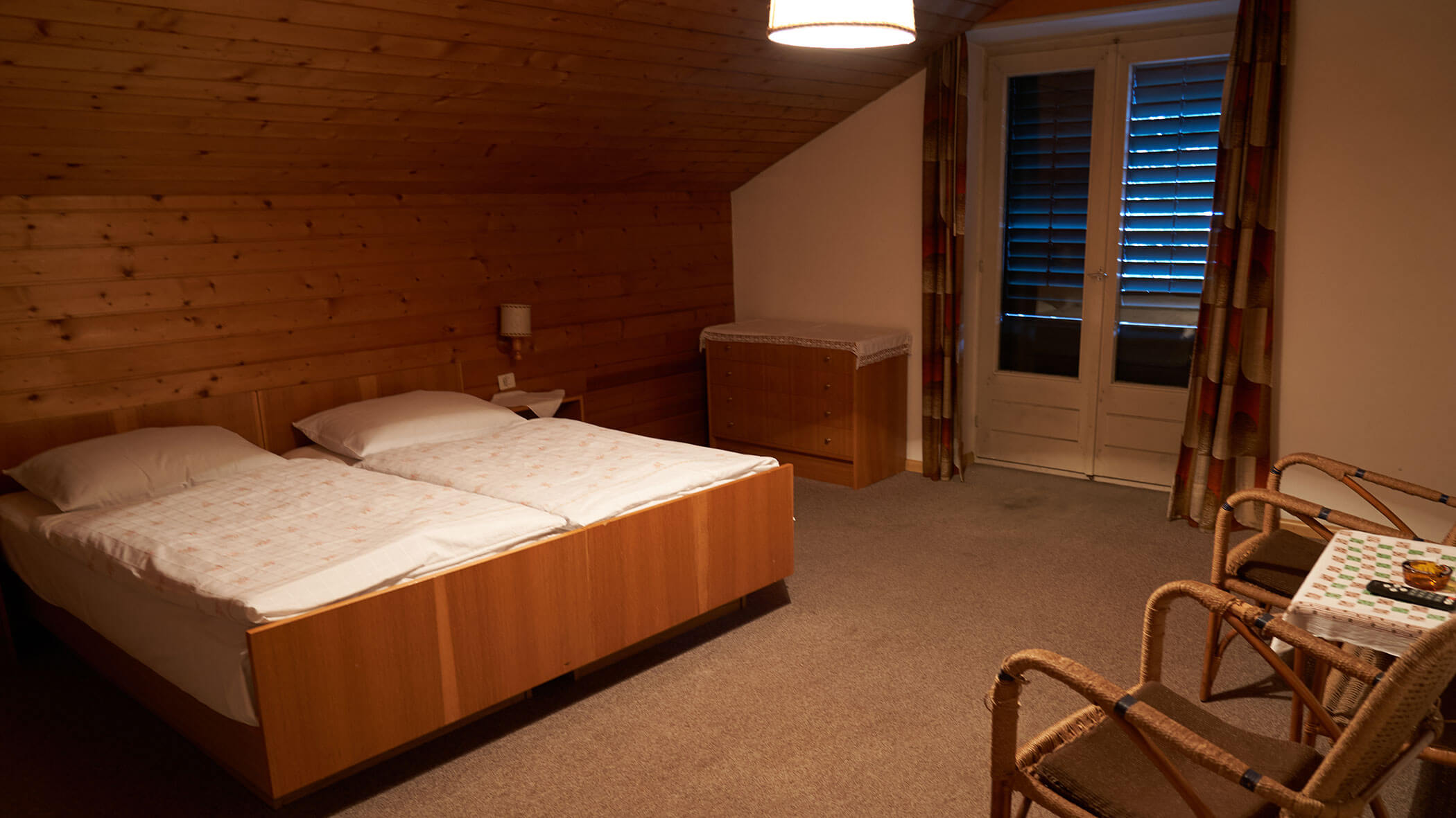 Cozy attic bedroom with twin beds, wooden walls, armchairs, and a table near glass doors with curtains. - Pension Pfarrwirt