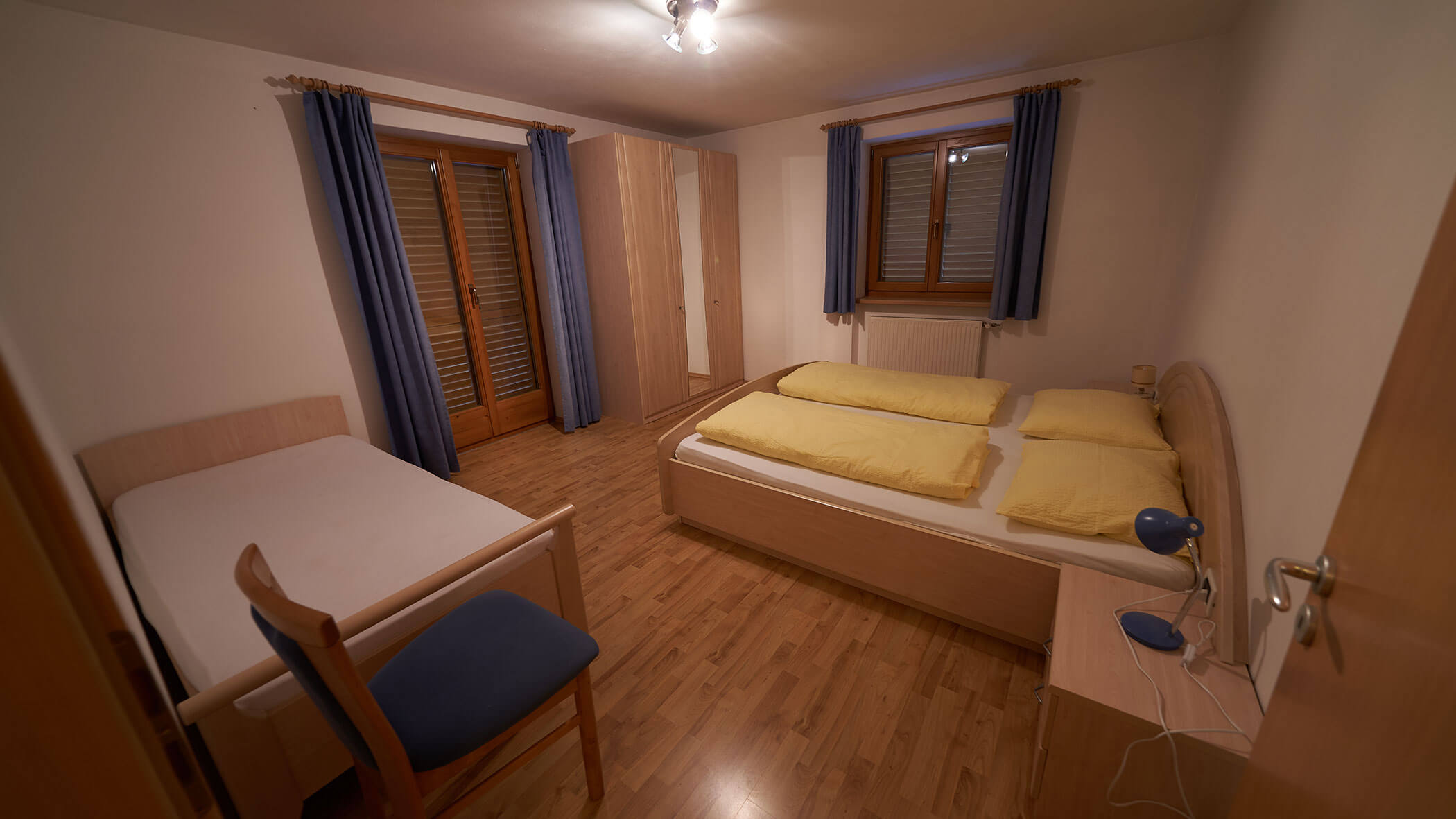 Bedroom with a double bed and a single bed, wooden floor, blue curtains, and yellow bedding. - Pension Pfarrwirt