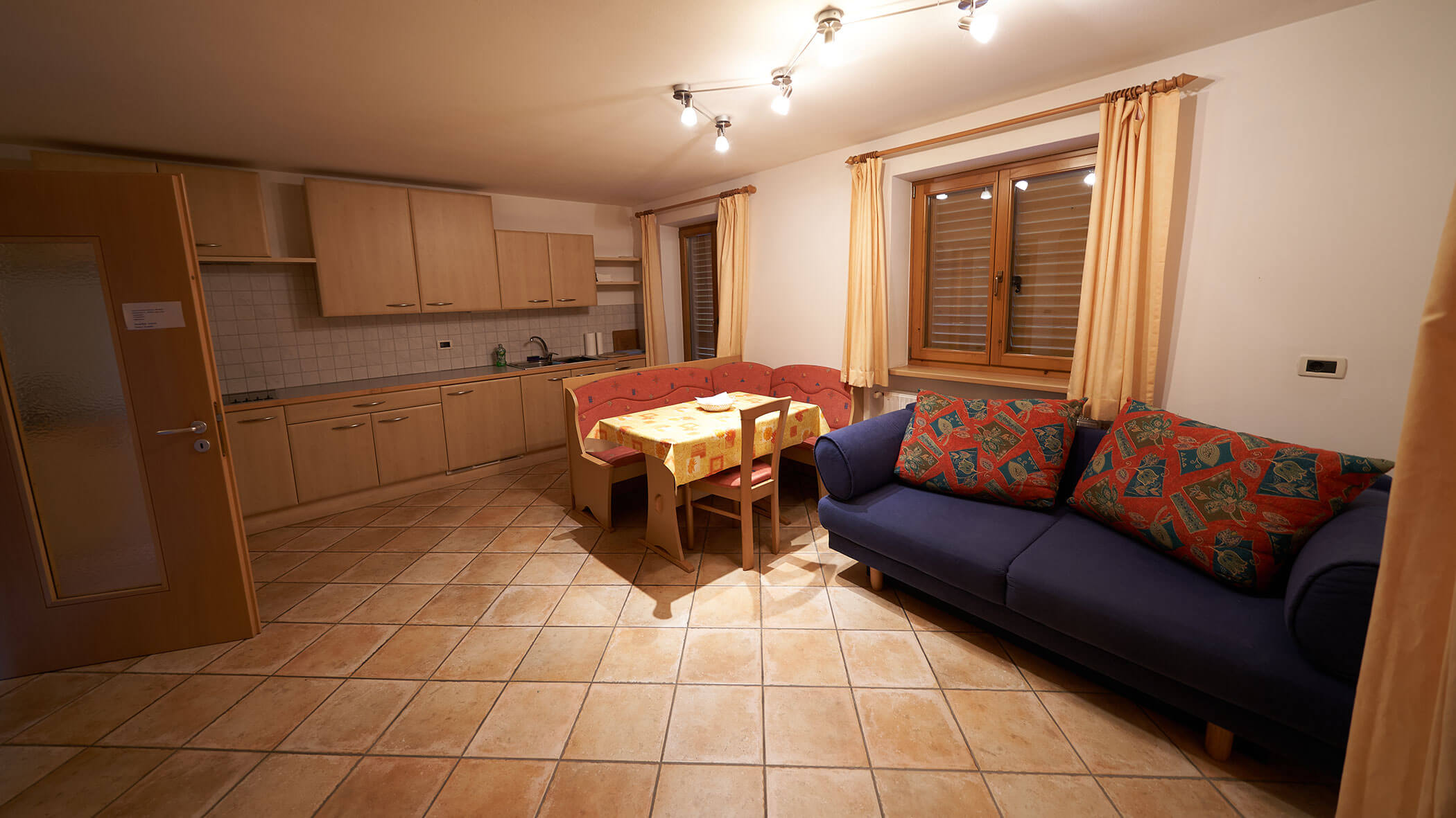 Cozy kitchen and living area with a blue sofa, dining table, wooden cabinets, and tiled floor. - Pension Pfarrwirt