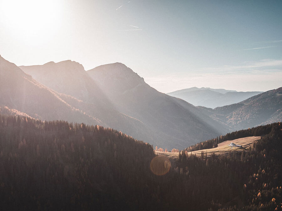 Sunlight shines over misty mountain peaks, forested slopes, and a small clearing with a house below. - Pension Pfarrwirt