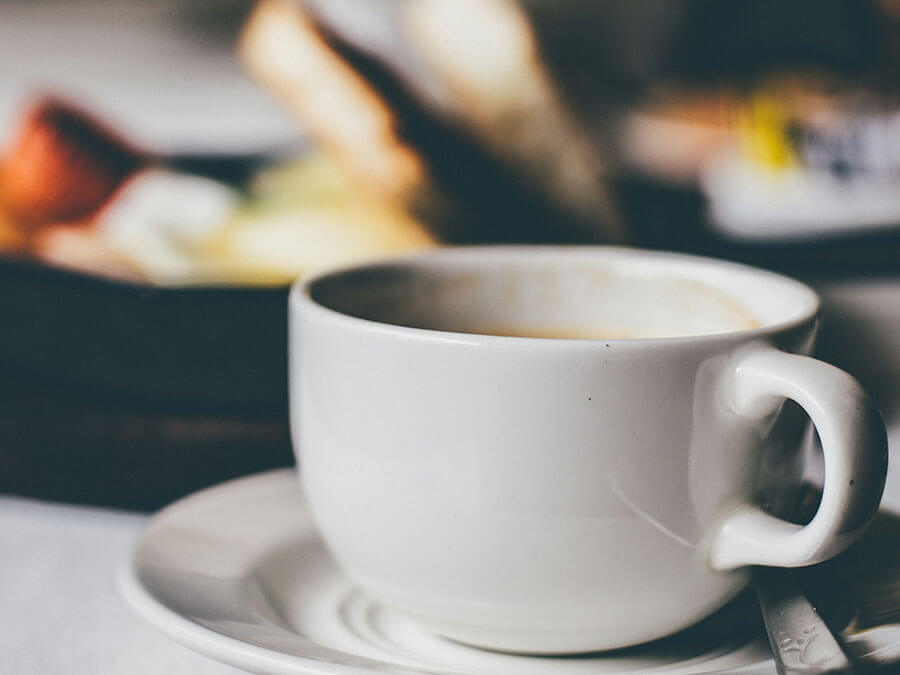 White coffee cup and saucer with a spoon, blurred breakfast food in the background. - Pension Pfarrwirt