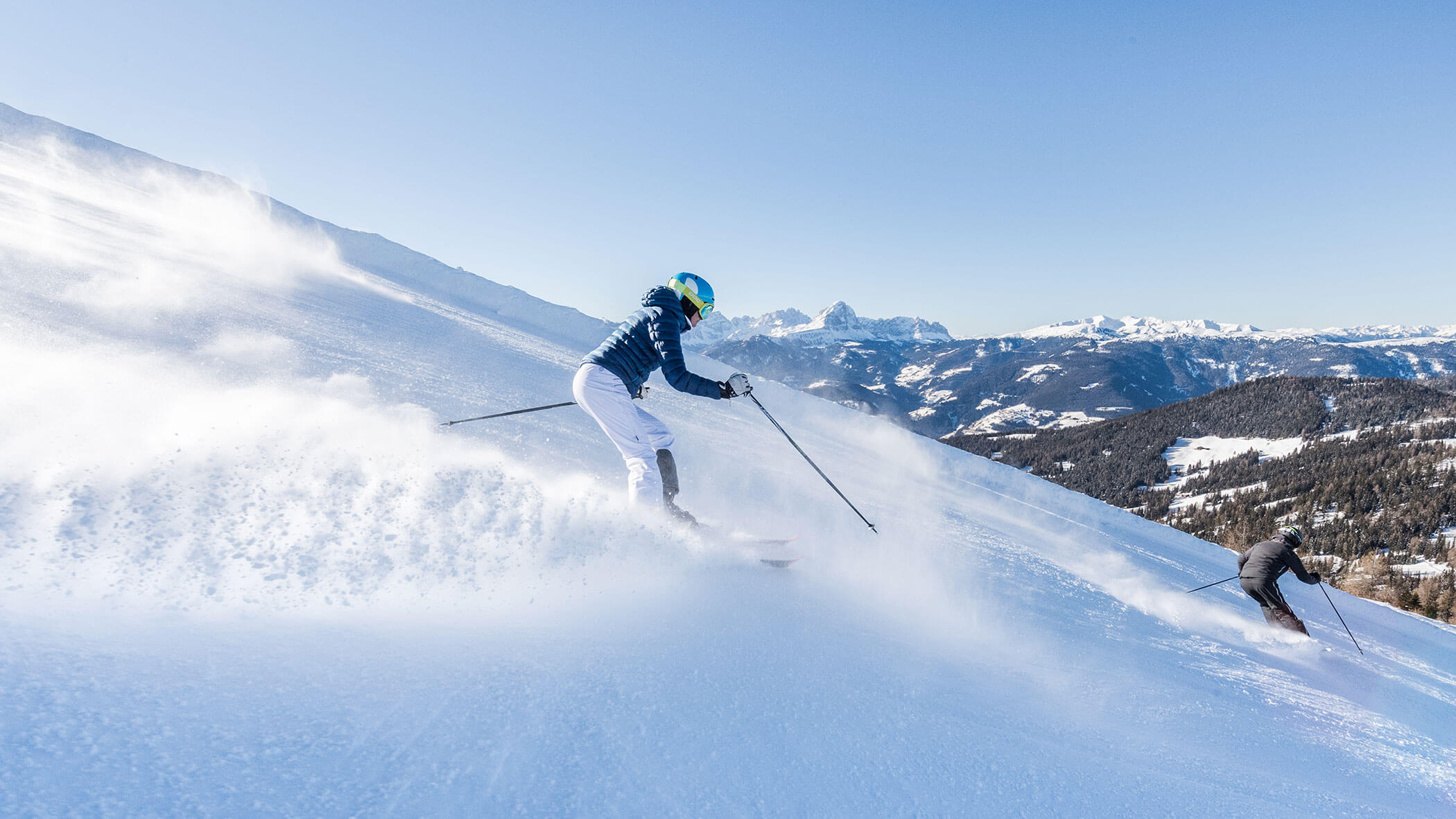 Two skiers glide down a snowy mountain slope under a clear blue sky, with distant peaks in the background. - Pension Pfarrwirt