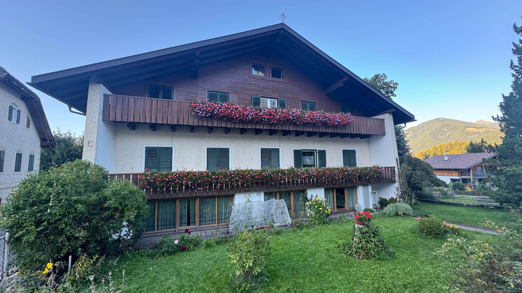 A large alpine-style house with flower boxes and a green lawn, set against mountains under a clear sky. - Pension Pfarrwirt