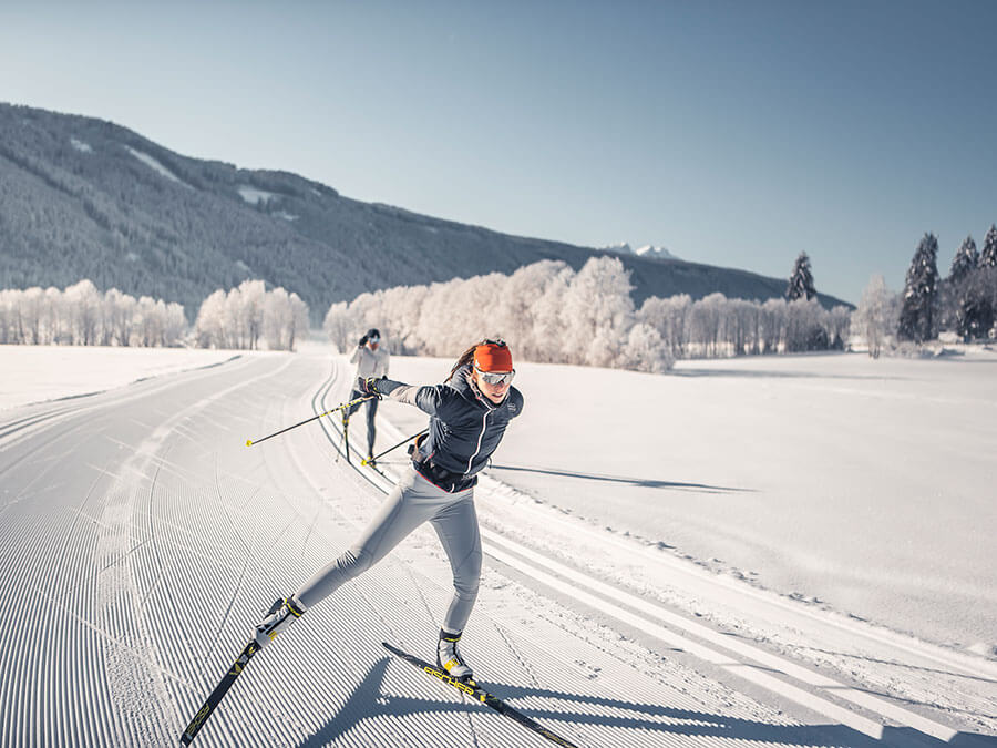 Two people cross-country skiing on a sunny, snowy trail with mountains and trees in the background. - Pension Pfarrwirt