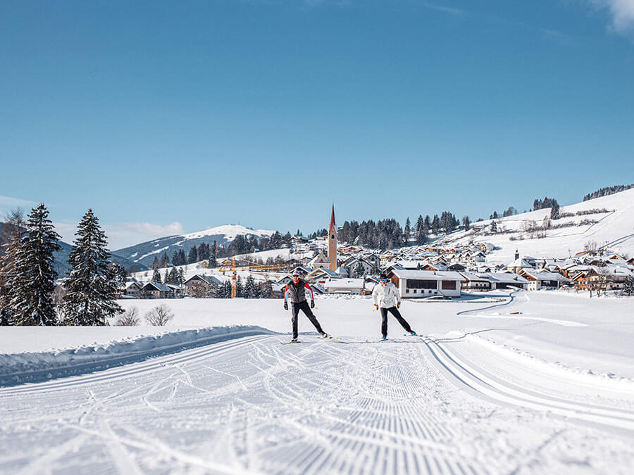 Two people cross-country skiing on snowy trails near a village with mountains and trees in the background. - Pension Pfarrwirt
