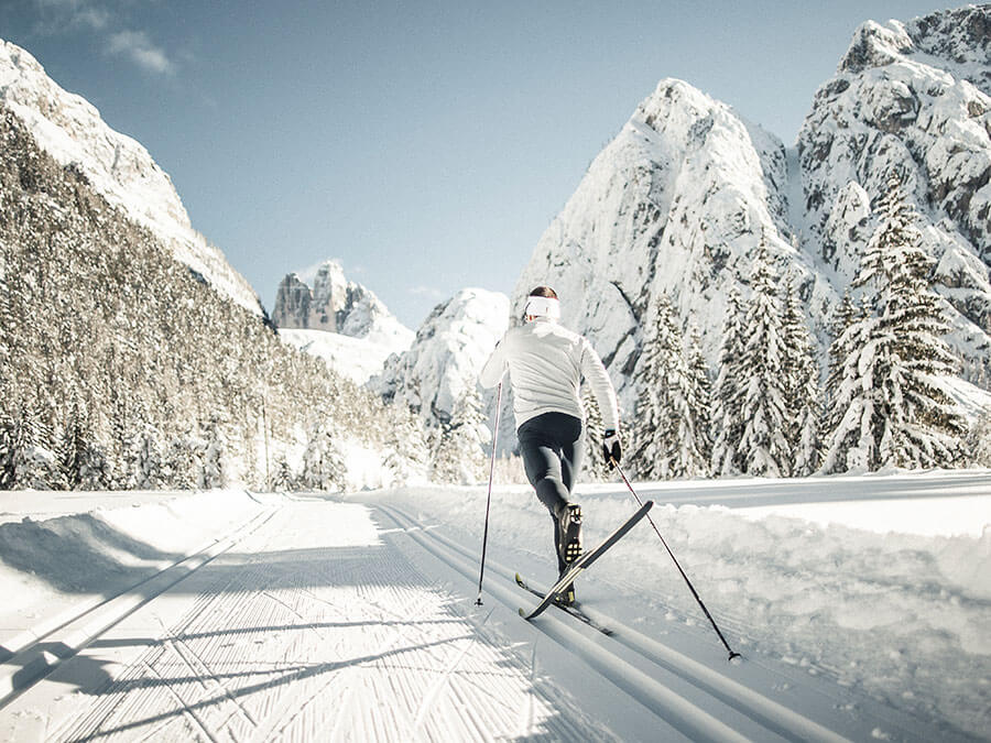 A person cross-country skiing on a snowy trail surrounded by snow-covered mountains and trees. - Pension Pfarrwirt