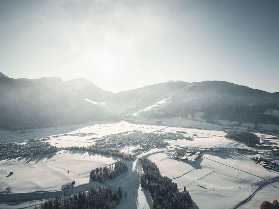 A snow-covered village nestled between mountains under a hazy, bright winter sky. - Pension Pfarrwirt
