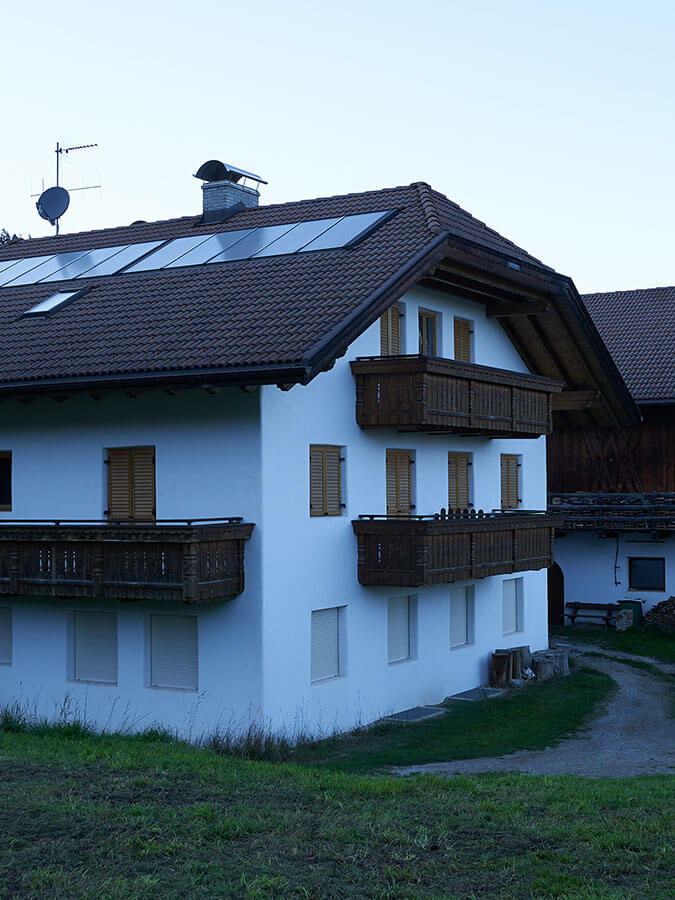 A white house with wooden balconies and closed shutters, set on a grassy area with a dirt path. - Pension Pfarrwirt