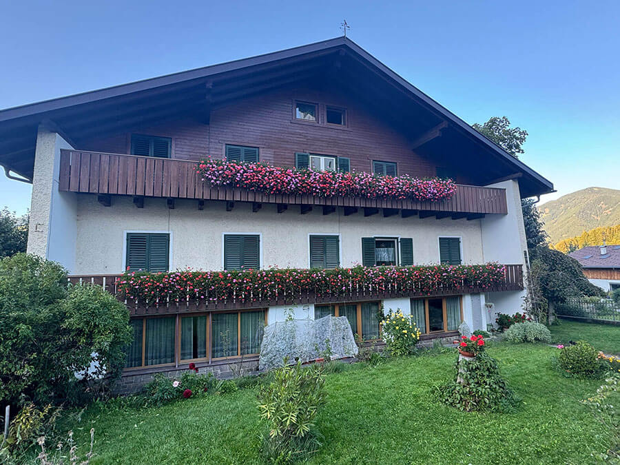 A house with balconies decorated with red flowers, green shutters, and a lawn with bushes and plants. - Pension Pfarrwirt