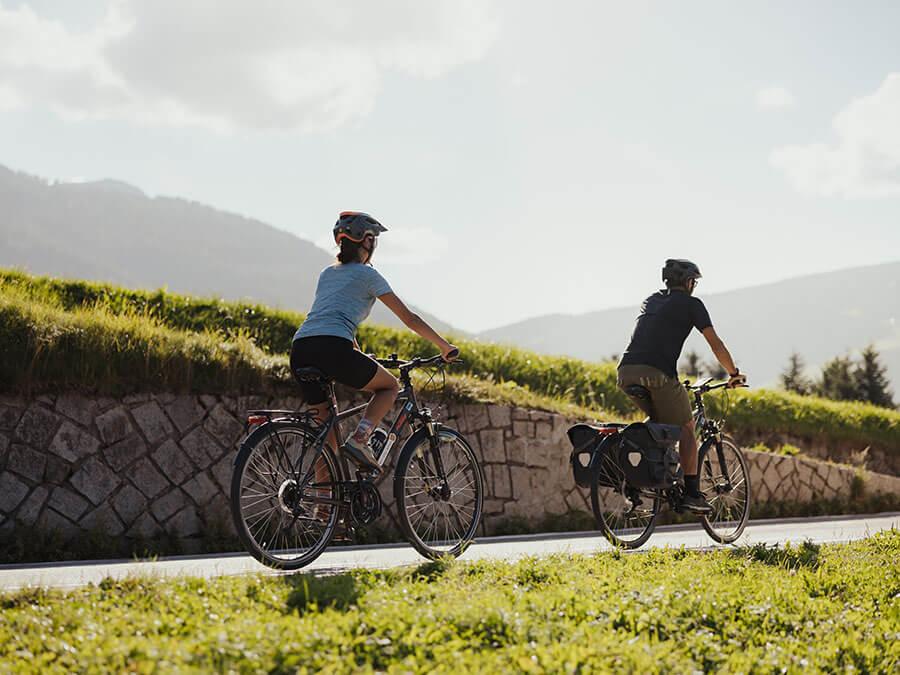 Two people wearing helmets ride bicycles on a rural road with grassy hills and mountains in the background. - Pension Pfarrwirt