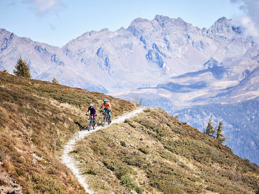 Two cyclists ride along a winding mountain trail with tall, rugged peaks in the background. - Pension Pfarrwirt