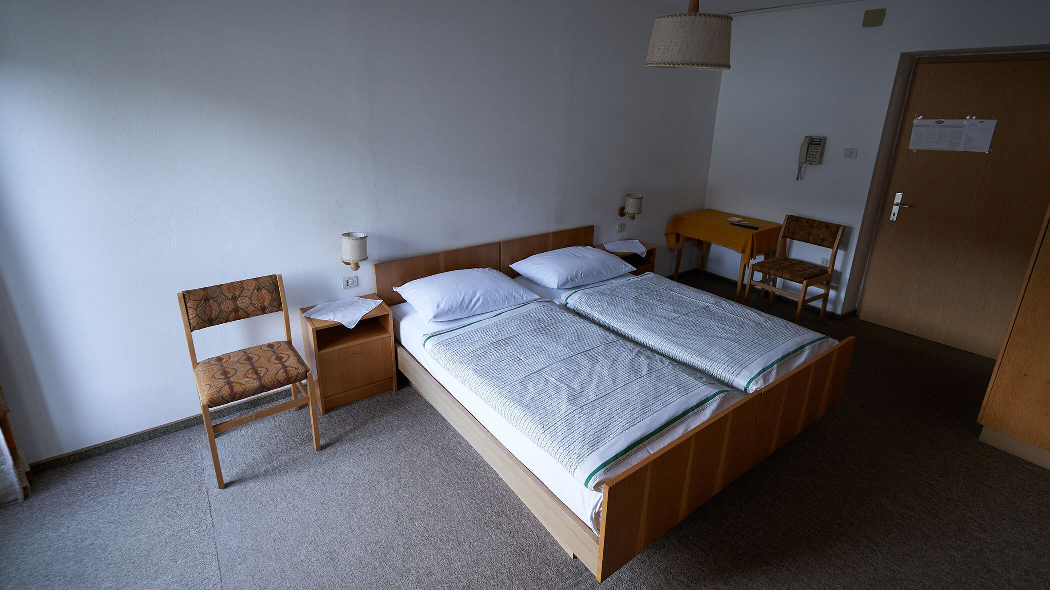 A small, simple hotel room with twin beds, two chairs, a desk, and a door. - Pension Pfarrwirt