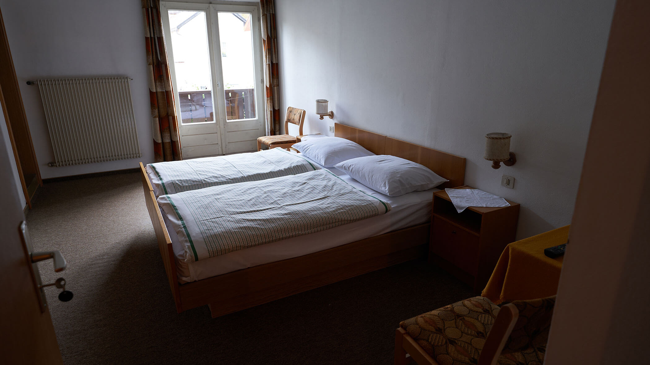 A simple hotel room with twin beds, a window with curtains, chairs, and a small table. - Pension Pfarrwirt