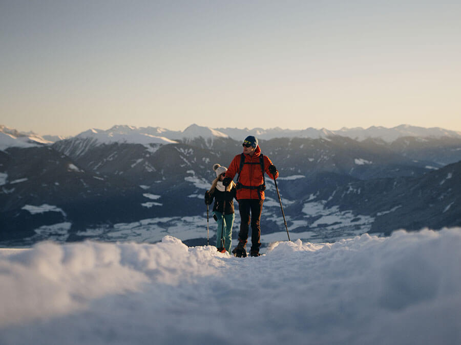 Two people hiking in snowy mountains at sunset, wearing winter gear and carrying hiking poles. - Pension Pfarrwirt