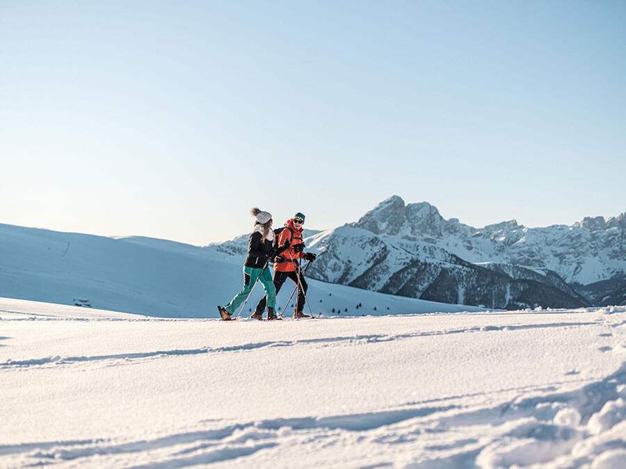 Two people snowshoeing on a snowy mountain slope with clear skies and distant peaks in the background. - Pension Pfarrwirt
