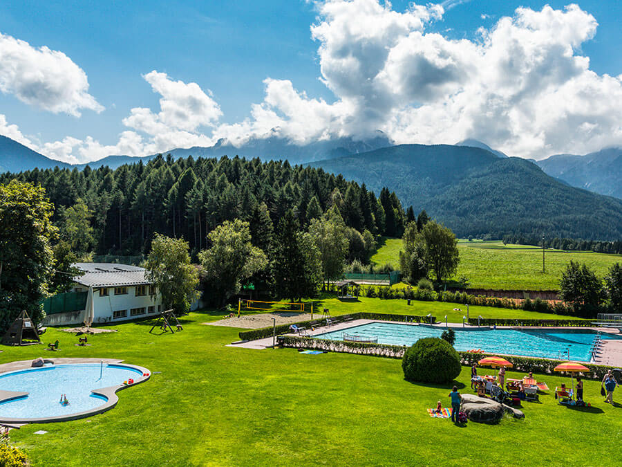 A pool in a grassy area with trees and mountains in the background. - Pension Pfarrwirt