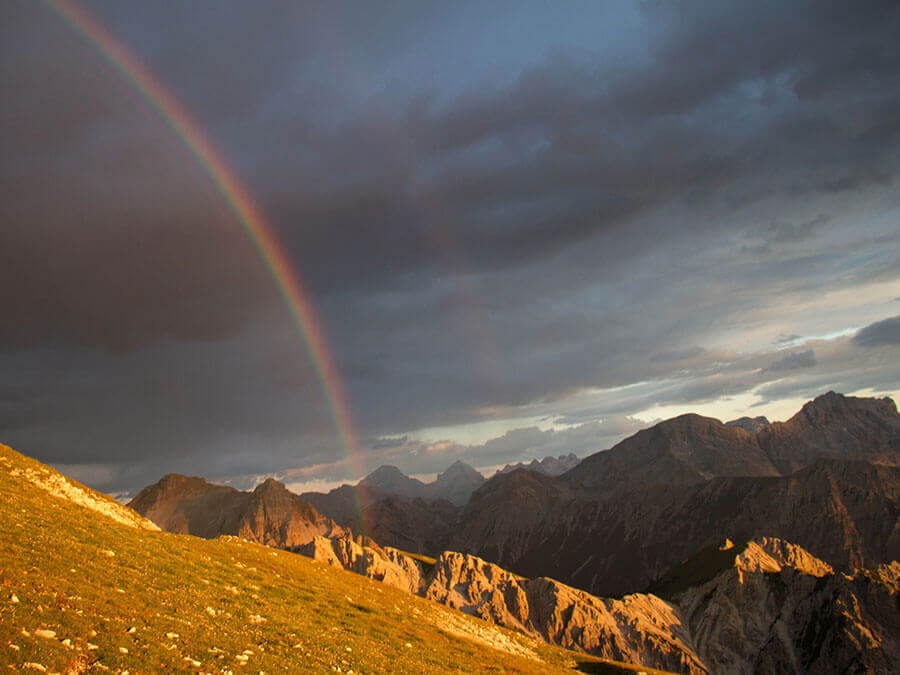 A rainbow arches over rugged mountains under a dramatic, cloudy sky at sunset. - Pension Pfarrwirt