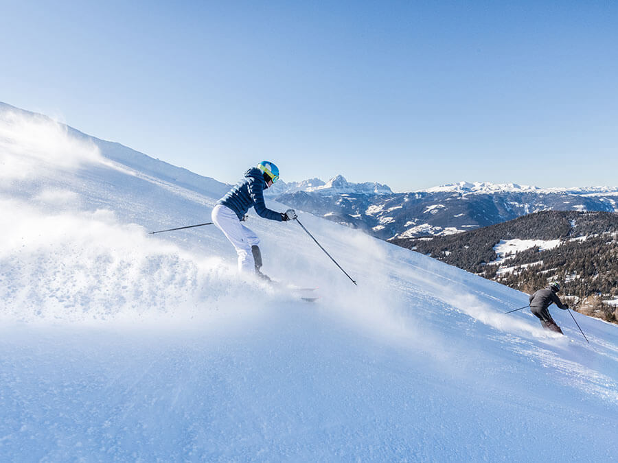 Two skiers in motion descend a snowy mountain slope under a clear blue sky with mountains in the background. - Pension Pfarrwirt