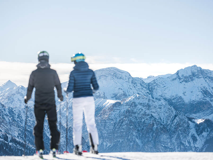 Two skiers stand on a snowy slope, looking at distant snow-covered mountains under a clear sky. - Pension Pfarrwirt