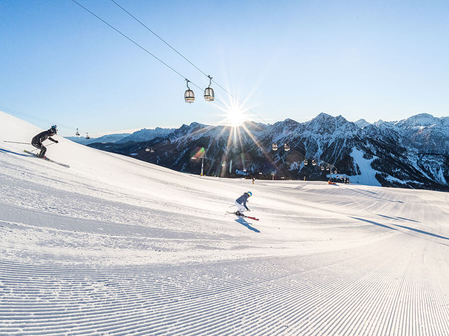 Two skiers descend a groomed slope with mountains and a ski lift in the background under a bright sun. - Pension Pfarrwirt