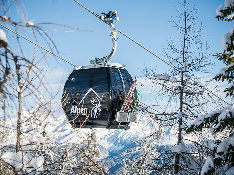 A gondola lift with skis attached glides above snowy trees and mountains under a clear blue sky. - Pension Pfarrwirt