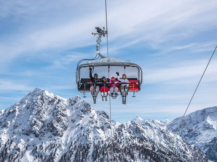 Four people ride a ski lift above snow-covered mountains under a blue sky with wispy clouds. - Pension Pfarrwirt