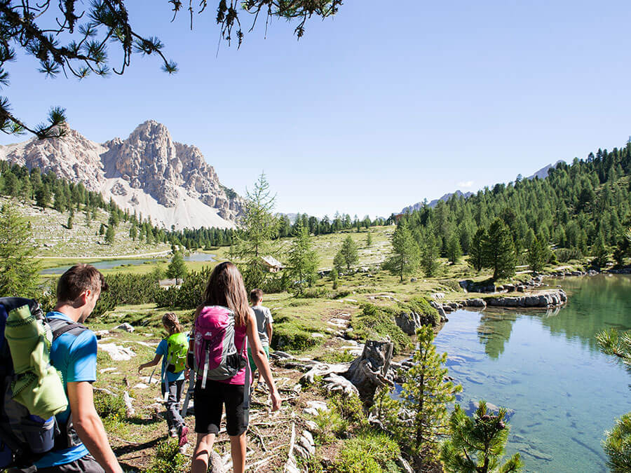 A group hikes along a scenic lakeside trail surrounded by mountains and trees under a clear blue sky. - Pension Pfarrwirt
