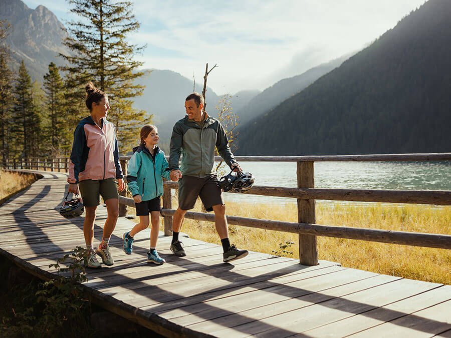 A family of three walks on a lakeside wooden path surrounded by mountains and trees, holding bike helmets. - Pension Pfarrwirt