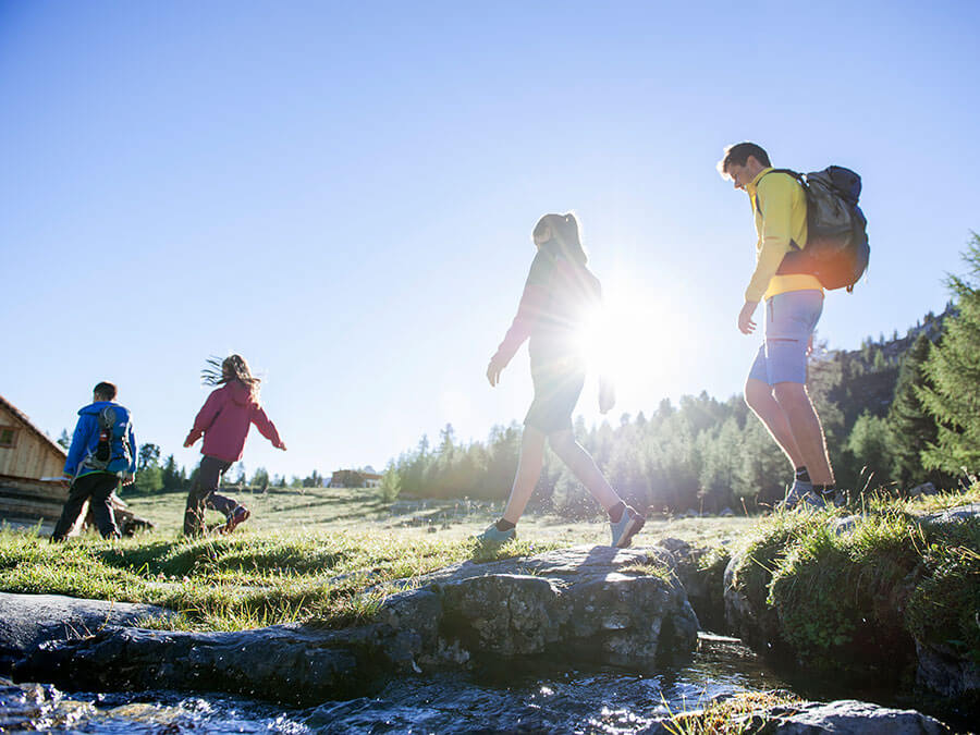 Four people hiking outdoors on a sunny day, crossing a small stream with trees and mountains in the background. - Pension Pfarrwirt