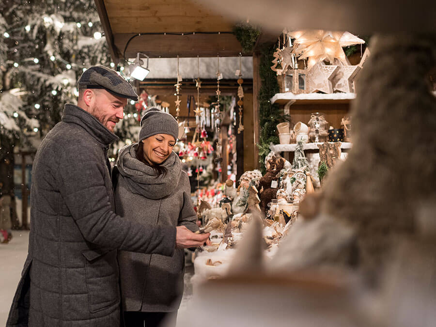 A couple in winter coats smile while browsing festive decorations at an outdoor holiday market. - Pension Pfarrwirt