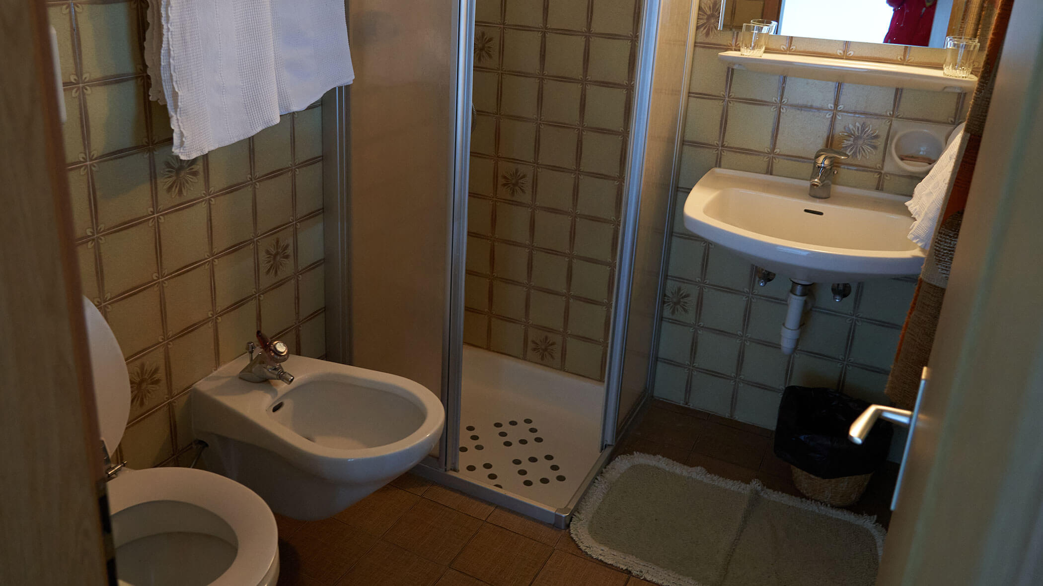Small bathroom with a toilet, bidet, shower, sink, towel, and greenish-brown tiled walls. - Pension Pfarrwirt